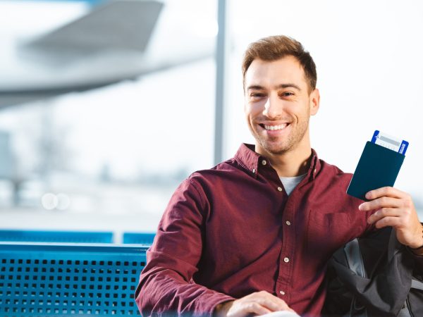cheerful man holding passport with air ticket in waiting hall of airport